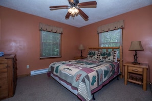Ein Schlafzimmer mit einem Bett, zwei Fenstern mit Jalousien, einem Deckenventilator, einem Nachttisch mit Lampe und einer Kommode.