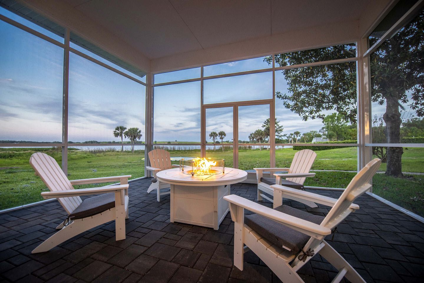 A covered outdoor seating area with a fire feature, surrounded by large windows offering a view of a grassy landscape.