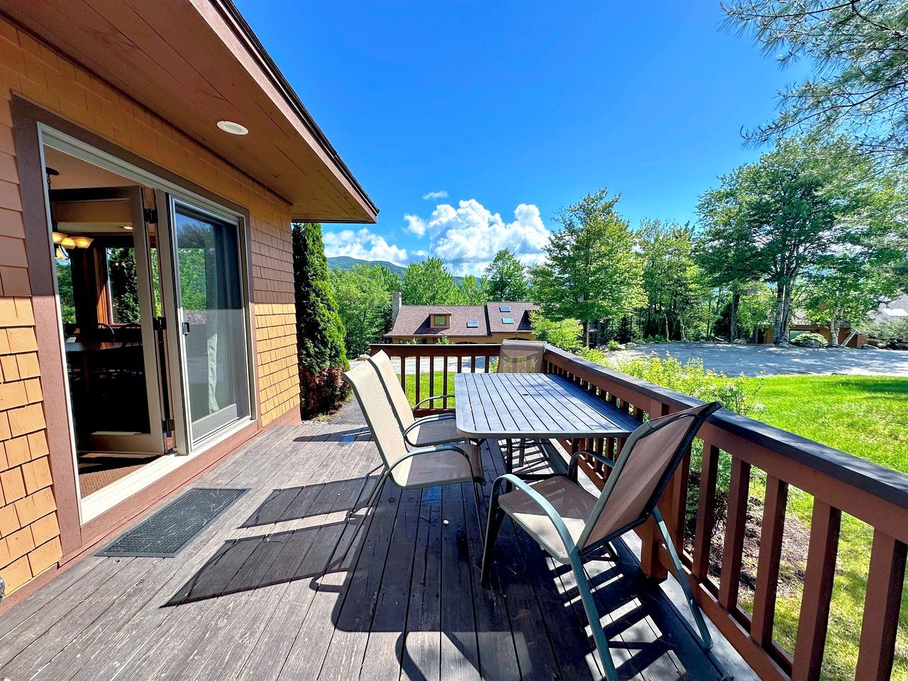 Une terrasse en bois avec une table et des chaises, une porte d’entrée en verre et une vue sur un paysage verdoyant.