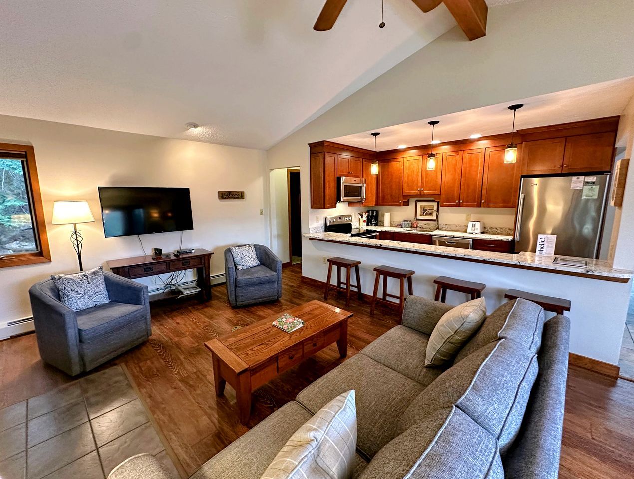 A modern living room with a sofa, armchairs, and a coffee table. A kitchen with wooden cabinets and stainless steel appliances is visible in the background.