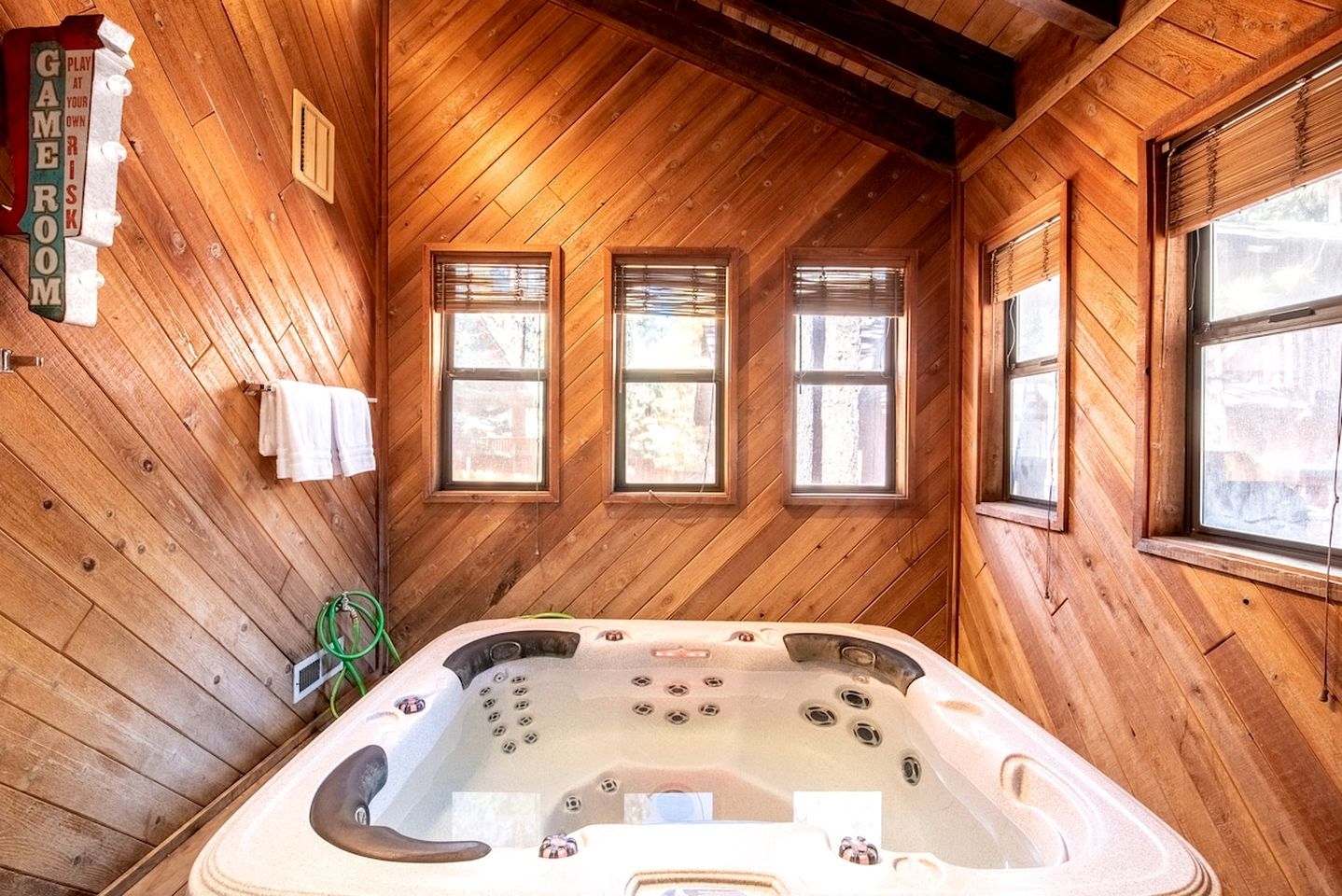 A wooden cabin interior with a hot tub, three windows, and a sign that reads 'GAMER ROOM.'