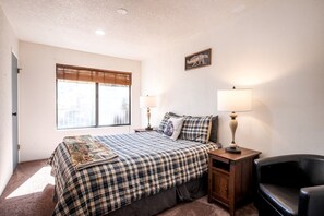 A bedroom with a bed, two bedside lamps, a chair, a window with blinds, and a wall-mounted sign. - Adventure Awaits: Big Bear Lake Mountain Cabin with Game Room + Hot Tub (Big Bear Lake)