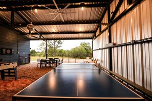 Mesa de pingue-pongue em ambiente interno com cadeiras, alvo de dardos e ventilador de teto em área coberta com vista para árvores.
