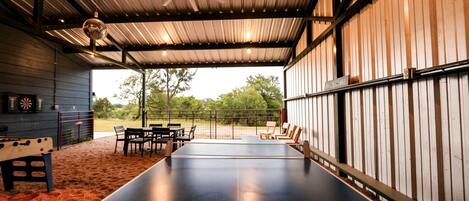 Una mesa de ping pong en el interior, con sillas, un dardo y un ventilador de techo en un espacio cubierto con vistas a árboles.