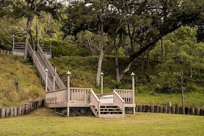 Eine hölzerne Treppe führt zu einer Plattform mit Geländern und Laternen, umgeben von Bäumen und grüner Landschaft.