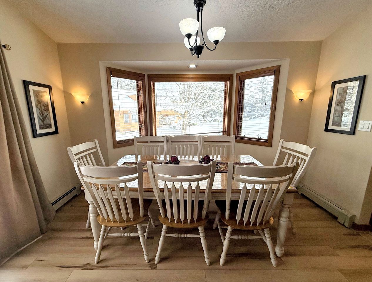 A dining area with a round table and six chairs, a chandelier, and large windows with blinds.