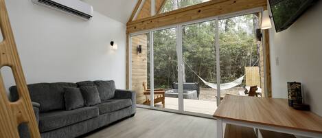 Un intérieur moderne avec un canapé gris, une table à manger en bois et de grandes portes vitrées donnant sur un espace extérieur agrémenté d’un hamac et d’un foyer.