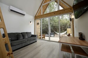 A modern interior with a grey sofa, wooden dining table, and large glass doors leading to an outdoor area with a hammock and a fire pit. - Luxury Cabin with Private Hot Tub - Cueta, Montgomery, TX (Montgomery)