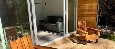 A wooden deck with two wooden chairs, a glass door leading to a room with a sofa and a fireplace, and a wooden ceiling.