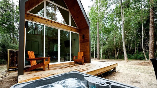 A wooden cabin with a hot tub, deck chairs, and a forest backdrop. - Luxury Cabin with Private Hot Tub - Cueta, Montgomery, TX (Montgomery)