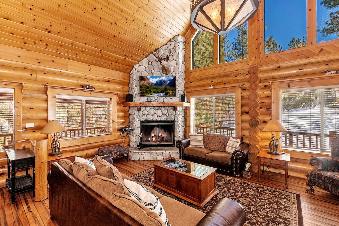 A log cabin interior with a stone fireplace, wooden furniture, and large windows.