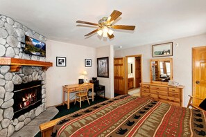 A bedroom with a stone fireplace, a bed with a patterned bedspread, a desk with a chair, a ceiling fan, and a dresser with a mirror. - Escape to Adventure: Mountain Cabin Holidays in Big Bear Lake (Big Bear Lake)