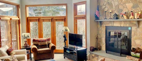 A spacious living room with a stone fireplace, large windows, and a television.