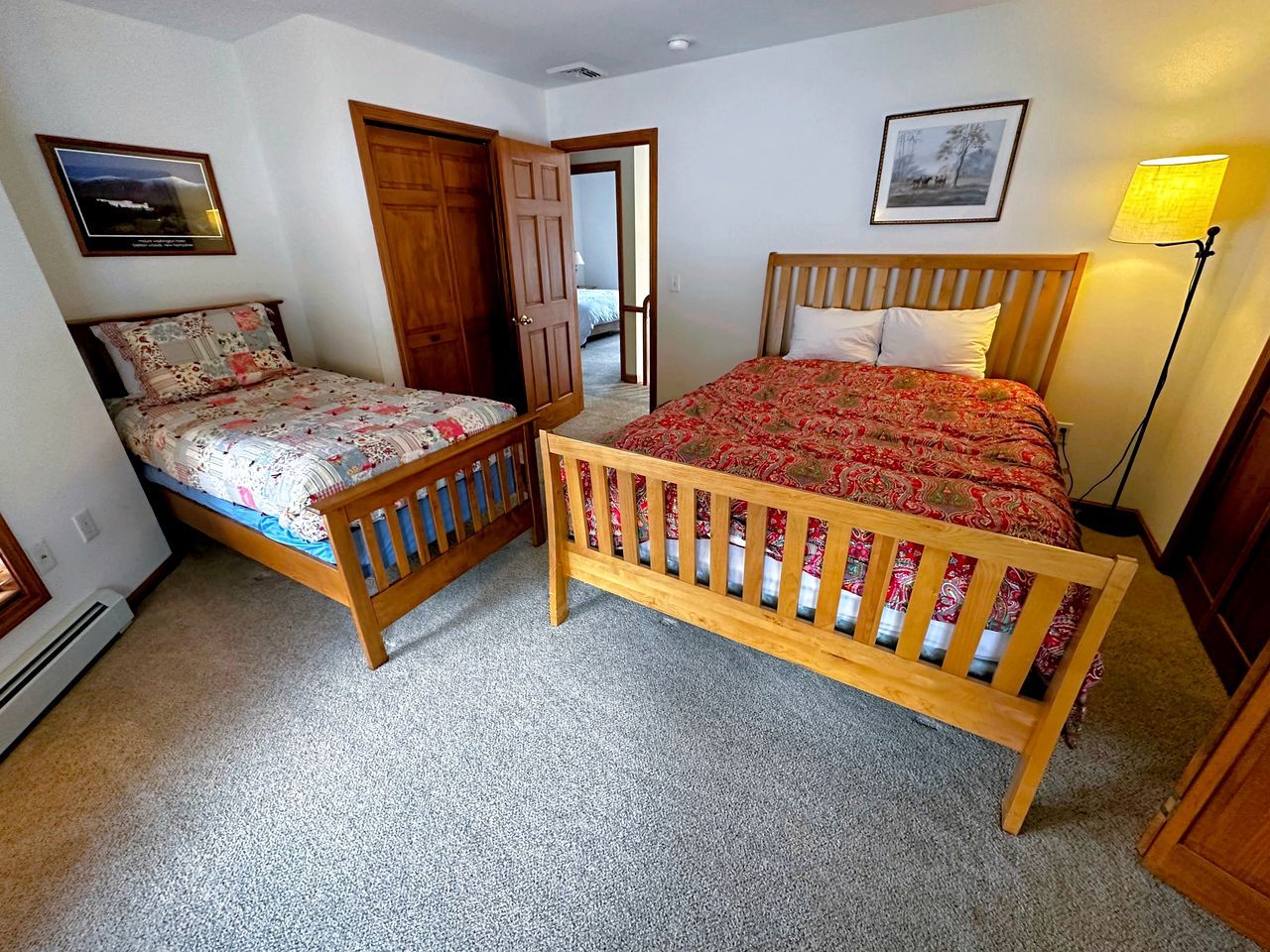 Un dormitorio con dos camas de madera, una puerta, un cuadro enmarcado en la pared y una lámpara.