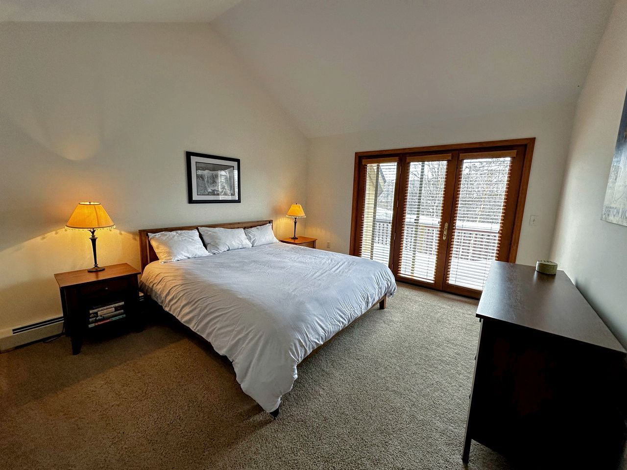 A bedroom with a bed, two bedside tables with lamps, a desk, a window with blinds, and a framed picture on the wall.