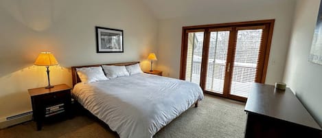 A bedroom with a bed, two bedside tables with lamps, a desk, a window with blinds, and a framed picture on the wall.