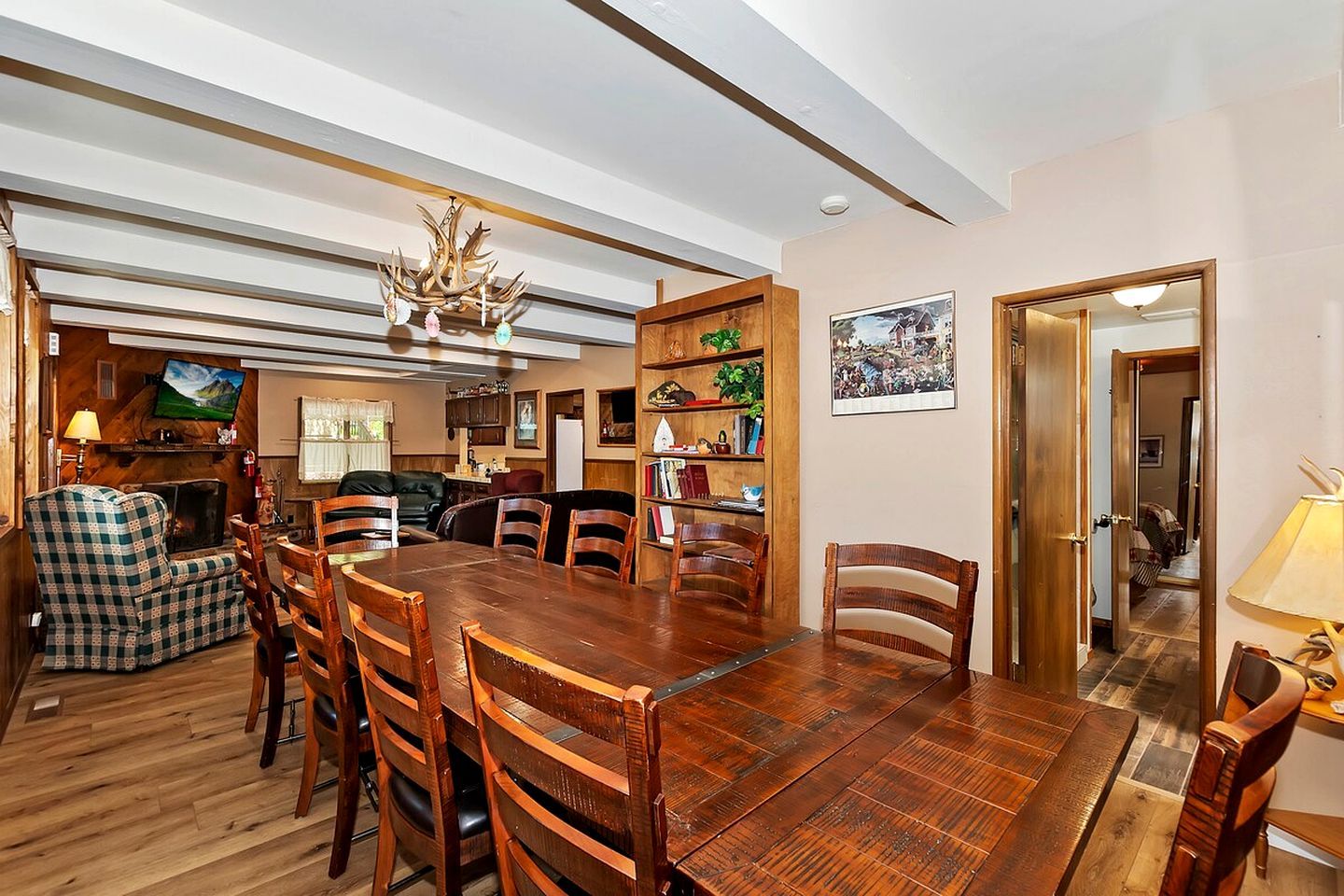 A dining area with a large wooden table, chairs, a chandelier, and a bookshelf with various items.