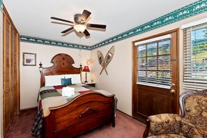 Quarto com cama de madeira, ventilador de teto, janela com persianas e cadeira com almofada estampada.