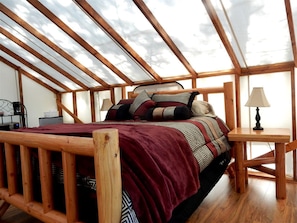 A bedroom with a wooden bed, a red blanket, patterned pillows, a nightstand, and a lamp.