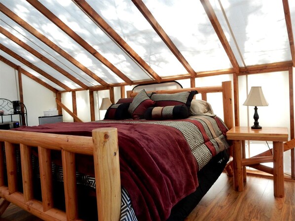 A bedroom with a wooden bed, a red blanket, patterned pillows, a nightstand, and a lamp.