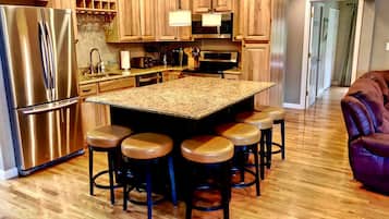 A kitchen with a central island, bar stools, stainless steel appliances, wooden cabinets, and a wooden floor.