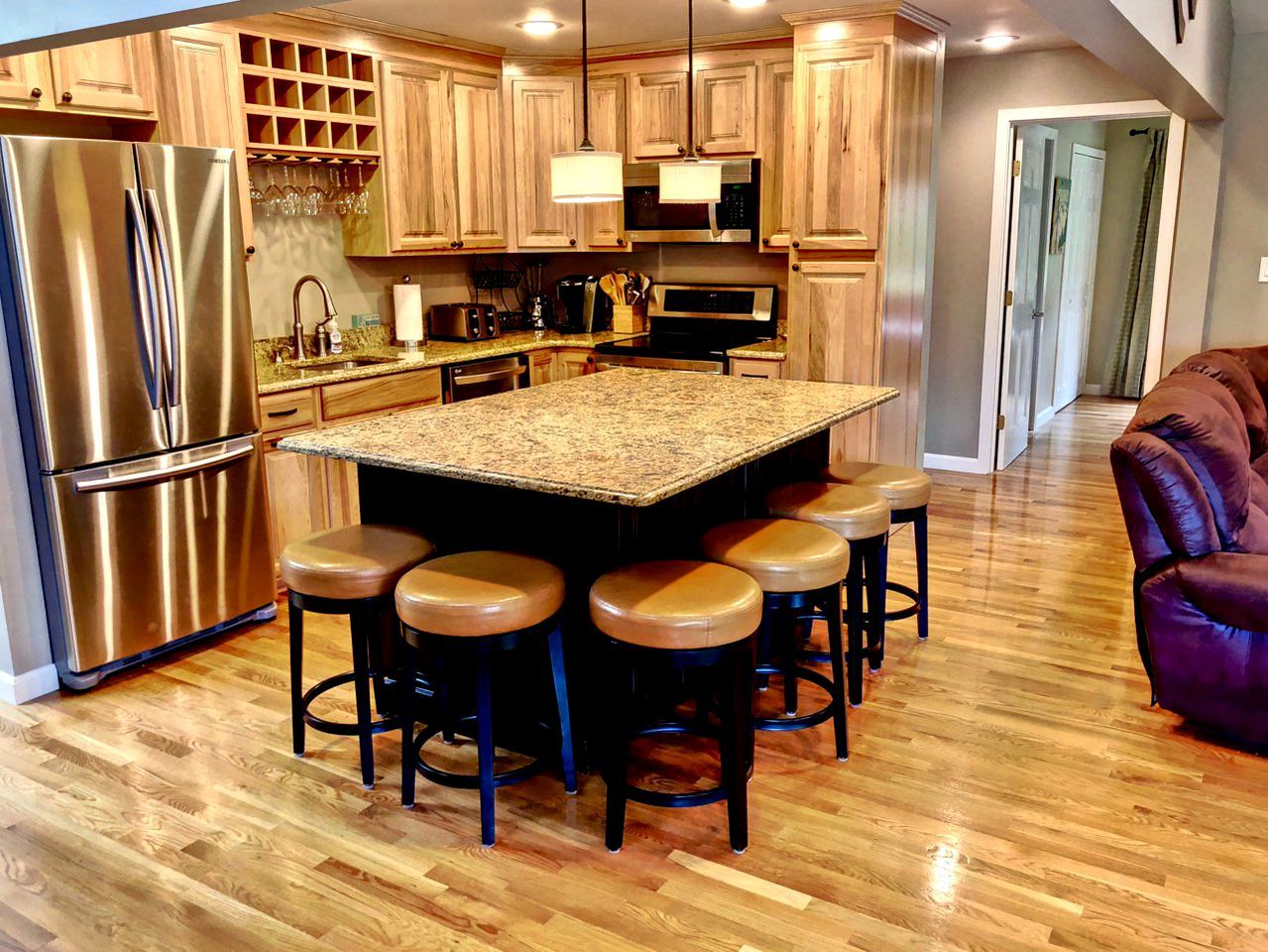 A kitchen with a central island, bar stools, stainless steel appliances, wooden cabinets, and a wooden floor.