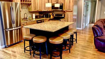 A kitchen with a central island, bar stools, stainless steel appliances, wooden cabinets, and a wooden floor.