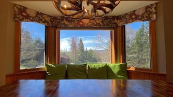 Un espace repas avec une table en bois et des sièges rembourrés de couleur verte, de grandes fenêtres offrant une vue sur les arbres et un luminaire au plafond au design original.