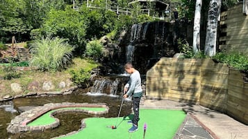 Une personne jouant au mini-golf près d’une petite cascade et de constructions à toits de chaume.