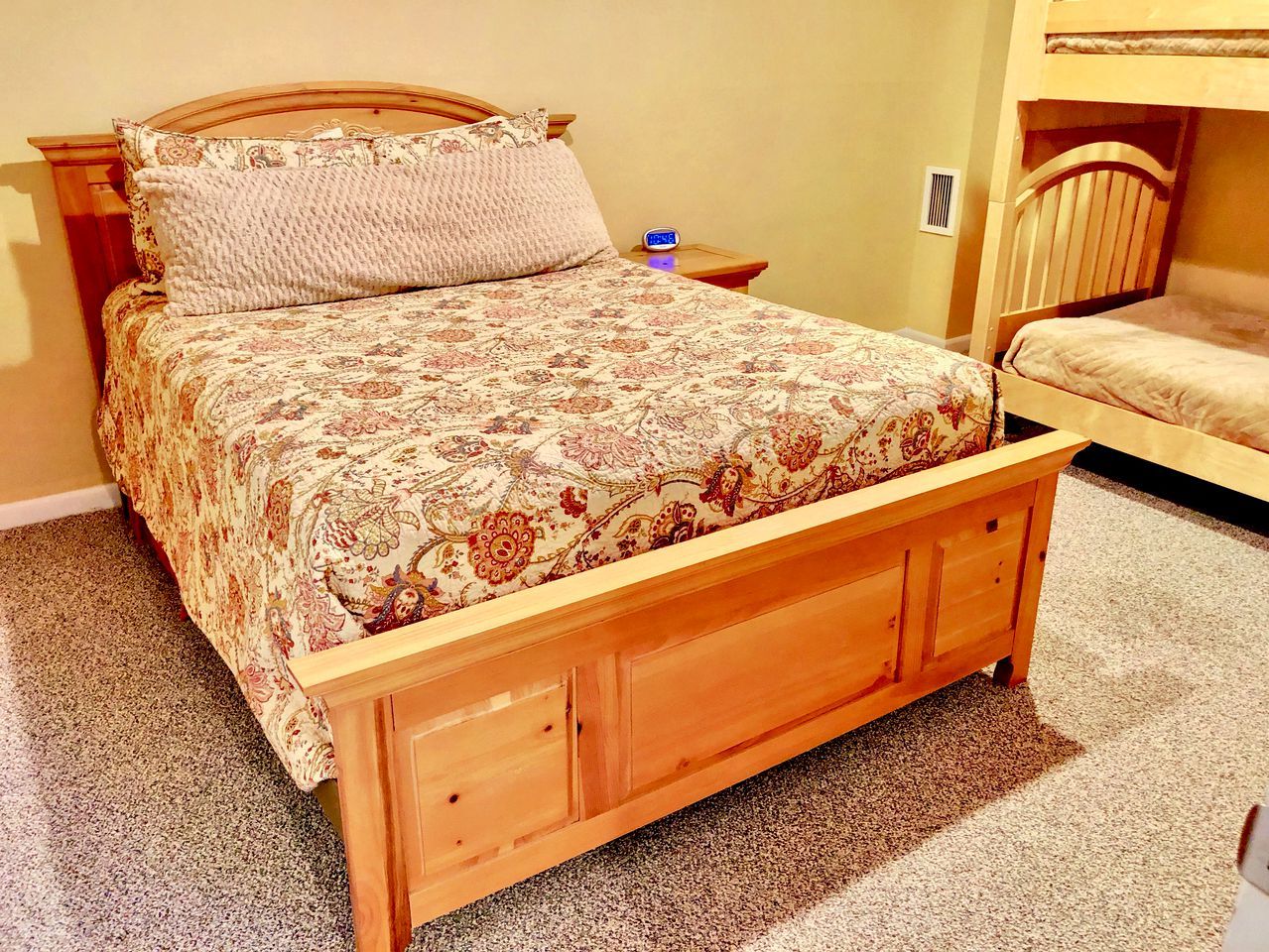 A wooden bed with a floral bedspread, a nightstand with a clock, and a bunk bed in the background.