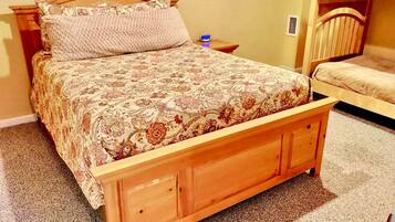A wooden bed with a floral bedspread, a nightstand with a clock, and a bunk bed in the background.