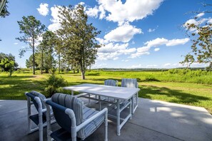 Um pátio externo com mesa e cadeiras, cercado por árvores e um gramado.