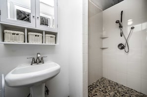 Un baño con lavamanos blanco, cestas de mimbre en repisas, ducha con cabezal de ducha manual y piso de ducha con diseño de guijarros.