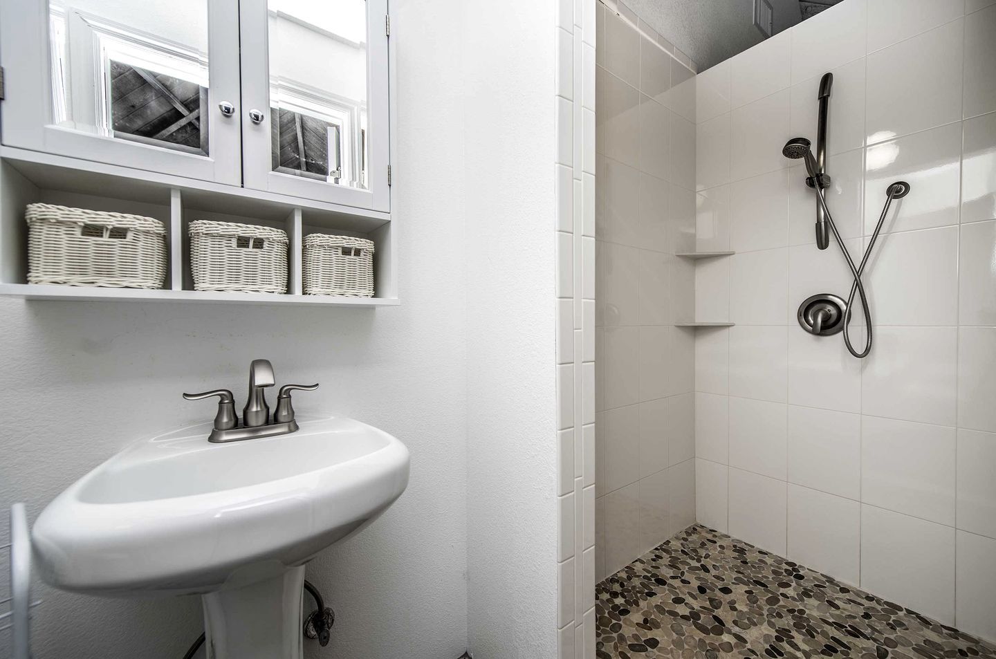 A bathroom with a white sink, wicker baskets on shelves, a shower with a handheld showerhead, and a pebble-patterned shower floor.