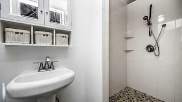 A bathroom with a white sink, wicker baskets on shelves, a shower with a handheld showerhead, and a pebble-patterned shower floor.