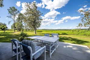 Um pátio externo com mesa e cadeiras, cercado por árvores e um gramado.