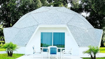 A geodesic dome structure with a patio area, outdoor seating, and a view of the surrounding greenery.