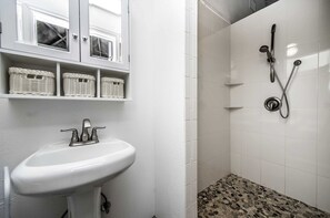 Casa de banho com lavatório branco, cestos de vime nas prateleiras, chuveiro com chuveiro manual e chão do chuveiro com padrão de pedras.
