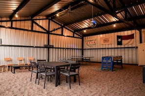 A spacious indoor area with a high ceiling, metal walls, and a table with chairs. There are flags and a sign on the walls. - Gorgeous Tiny House with Fire Pit, Games and Barbecue in San Saba, Texas (San Saba)