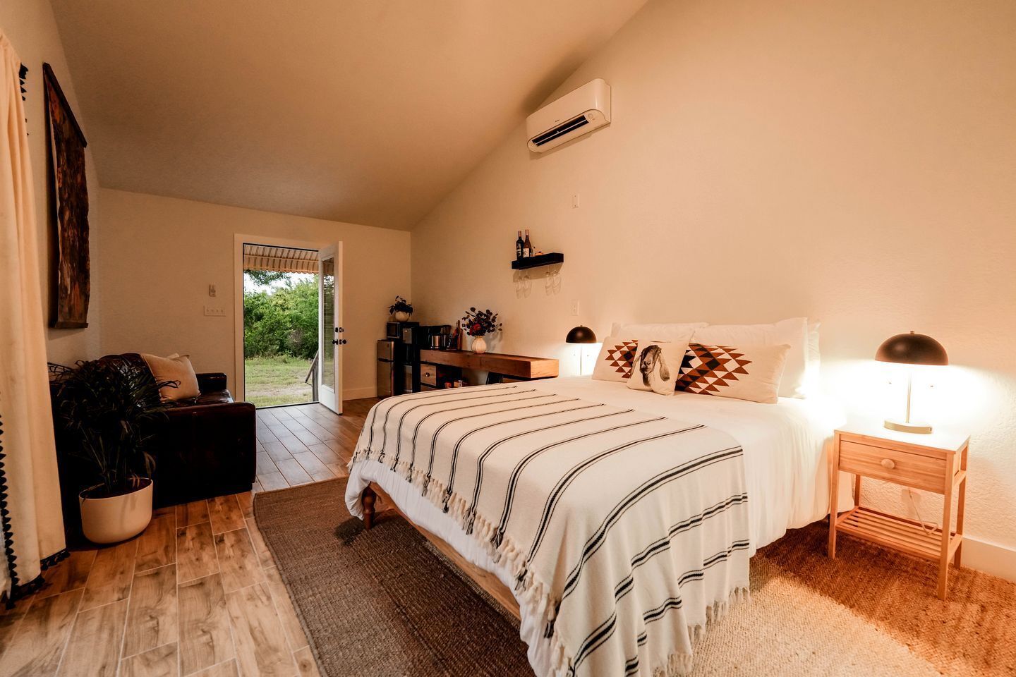 A bedroom with a bed, bedside table, lamp, and a view of the outdoors.