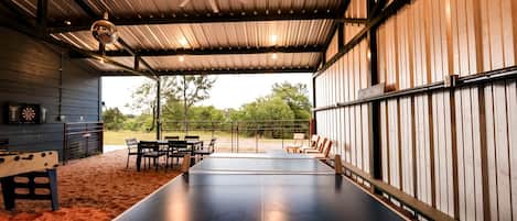 Una mesa de ping pong en el interior, con sillas, un dardo y un ventilador de techo en un espacio cubierto con vistas a árboles.