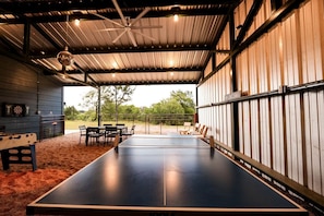 An indoor ping pong table with chairs, a dartboard, and a ceiling fan in a covered area with a view of trees. - Gorgeous Tiny House with Fire Pit, Games and Barbecue in San Saba, Texas (San Saba)