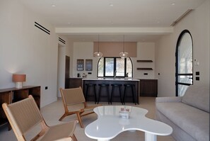 Ein modernes Wohnzimmer mit einem weißen Couchtisch, zwei Korbstühlen, einem grauen Sofa und einer Küchenzeile mit Barhocker und Pendelleuchten.