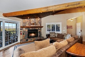A living room with a stone fireplace, a wooden ceiling beam, a large sofa, and a view through glass doors. - Secluded Cabin with Breathtaking Lake Views and Foosball Table in Big Bear Lake, California (Big Bear Lake)