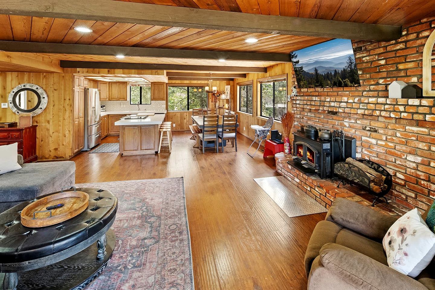 A spacious living area with a fireplace, wooden flooring, and a kitchen in the background.