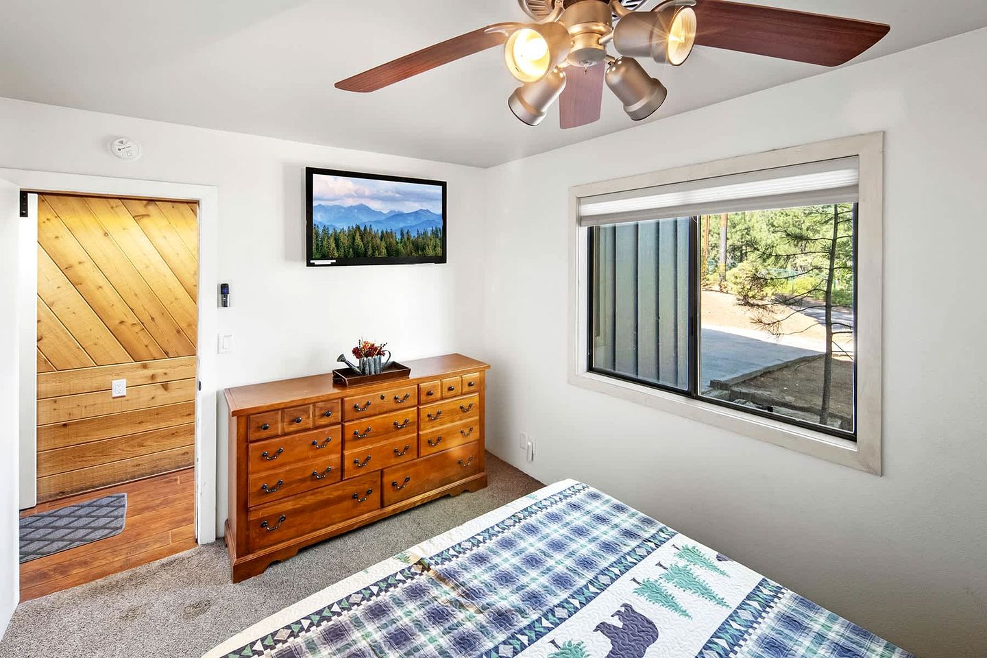 Un dormitorio con cama, cómoda de madera, ventilador de techo, ventana con vista a árboles y puerta con diseño de panel de madera.