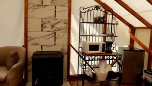 A cozy attic room with a fireplace, a sofa, a metal shelf with kitchen appliances, and a wooden ceiling.