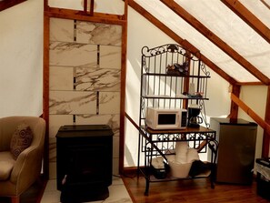 A cozy attic room with a fireplace, a sofa, a metal shelf with kitchen appliances, and a wooden ceiling.