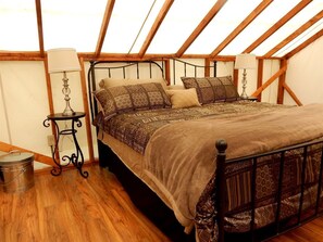 A bedroom with a wooden ceiling, a metal bed frame, patterned bedding, a small table with a lamp, and a trash can.