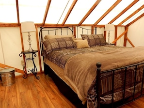 Un dormitorio con techo de madera, estructura de cama de metal, ropa de cama estampada, una mesita con una lámpara y un cubo de basura.